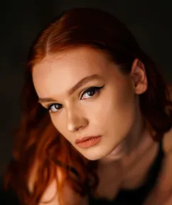 Portrait of a red-haired woman with winged eyeliner and neutral makeup against a dark background.